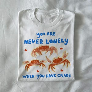 May include: White crew neck t-shirt with a crab graphic in orange and blue. The text reads "YOU ARE NEVER LONELY WHEN YOU HAVE CRABS". The design features three crabs and small red stars.