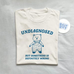 Op de afbeelding: Een wit T-shirt met blauwe tekst die "Undiagnosed but something is definitely wrong" luidt. Een cartoonbeer met een overall is op het shirt gedrukt.