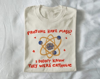 May include: Cream-colored t-shirt with a science-themed graphic. The design features an atom model in orange and blue, with the text "PROTONS HAVE MASS? I DIDN'T KNOW THEY WERE CATHOLIC" in red. The shirt is folded.