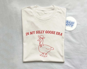 May include: A white t-shirt featuring a red outline of a goose wearing a cowboy hat and boots. The text "IN MY SILLY GOOSE ERA" is printed in red on the shirt.
