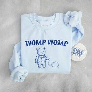 Può includere: Felpa blu chiaro con un orso dei cartoni animati che tiene un palloncino e il testo "WOMP WOMP". La felpa ha un collo rotondo e maniche lunghe. Un adesivo bianco con il testo "SILLY CITY" è sullo sfondo.