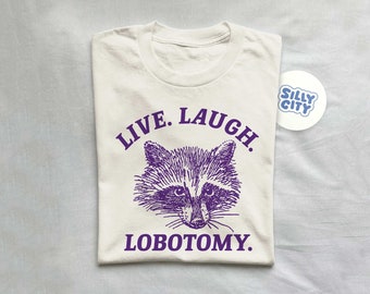 May include: A white t-shirt with purple text that reads "Live. Laugh. Lobotomy." and a purple illustration of a raccoon's face.