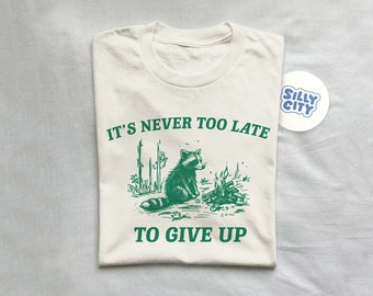 May include: A white t-shirt with a green graphic of a raccoon sitting by a campfire. The text "It's never too late to give up" is printed in green on the shirt.