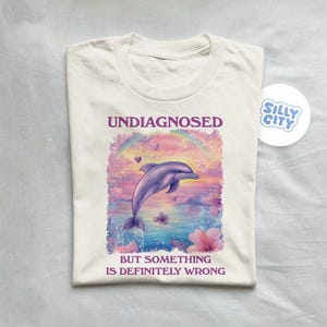 Op de afbeelding: Wit T-shirt met een afbeelding van een dolfijn die over een regenboog met roze bloemen springt. De tekst "Undiagnosed but something is definitely wrong" staat op het shirt gedrukt.