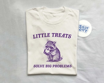 Op de afbeelding: Een wit T-shirt met een paarse afbeelding van een wasbeer die een traktatie eet. De tekst "Little Treats Solve Big Problems" is in paars onder de wasbeer gedrukt.