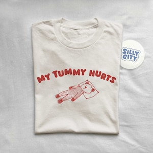 May include: A white t-shirt with the text "My tummy hurts" in red lettering. A cartoon illustration of a bear lying down with a pillow is below the text. A sticker with the text "Silly City" in blue lettering is in the top right corner of the image.