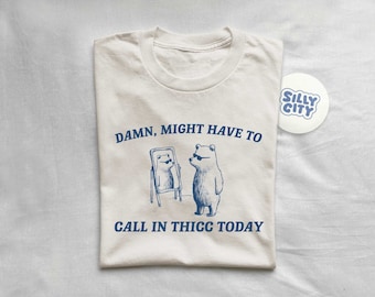 Op de afbeelding: Een wit T-shirt met een blauwe afbeelding van een beer met een zonnebril die in een spiegel kijkt. De tekst "DAMN, MIGHT HAVE TO CALL IN THICC TODAY" staat op het shirt gedrukt.