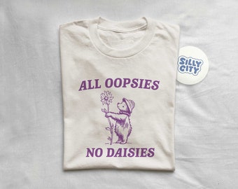 May include: A beige t-shirt with a purple graphic of a bear holding a flower. The text "ALL OOPSIES NO DAISIES" is printed on the shirt.