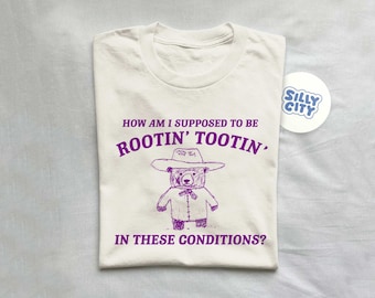 May include: A white t-shirt with purple text that reads "HOW AM I SUPPOSED TO BE ROOTIN' TOOTIN' IN THESE CONDITIONS?" and a cartoon drawing of a bear wearing a cowboy hat.