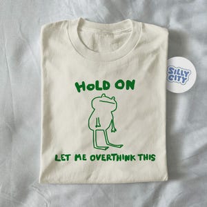 Può includere: T-shirt color crema con illustrazione di rana verde e la scritta "HOLD ON LET ME OVERTHINK THIS." Un adesivo rotondo bianco con "SILLY CITY" in blu è accanto alla maglietta piegata.