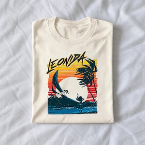 May include: A cream-colored t-shirt featuring a graphic print of a sunset over the ocean with palm trees, a sailboat, and a person standing on the shore. The text LEONIDA is printed in a bold, black, stylized font above the sunset scene.