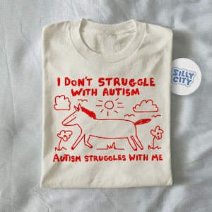 Puede incluir: Camiseta color crema con texto rojo y un dibujo de un caballo. El texto dice "I DON'T STRUGGLE WITH AUTISM, AUTISM STRUGGLES WITH ME." Una pegatina "Silly City" es visible. La camiseta está doblada.