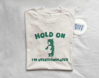 以下が含まれることがあります： 緑色のクマのイラストと「HOLD ON I'M OVERSTIMULATED」の文字が入ったクリーム色のTシャツ。青色の「SILLY CITY」と書かれた白い丸いステッカーがTシャツの横にあります。Tシャツは白い表面に折りたたまれています。