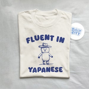 Könnte beinhalten: Ein weißes T-Shirt mit blauem Text, der "Fluent in Japanese" lautet, und einer Cartoon-Zeichnung eines Bären mit Cowboyhut. Das Shirt hat auch einen blauen Aufkleber mit der Aufschrift "Silly City".