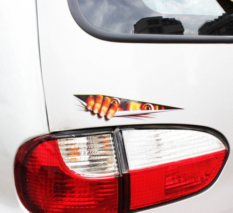 Puede incluir: Un coche blanco con una pegatina de una mano que sale de detr&aacute;s de un trozo de papel rasgado. La mano tiene una expresi&oacute;n espeluznante con un ojo y una boca visibles. El coche tiene una luz trasera roja.