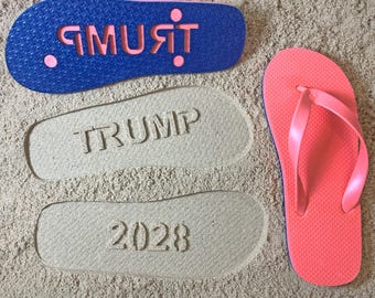 Może przedstawiać: Para koralowych klapek z niebieskimi podeszwami i różowymi paskami obok niebieskiej podeszwy klapek z wyciętym różowym napisem "TRUMP". Odbicia stóp w piasku z napisami "TRUMP" i "2028".