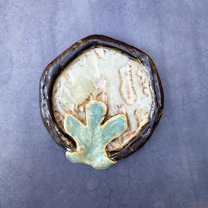 May include: A handmade ceramic dish with a dark brown rim and a light green, beige, and brown interior. A light green leaf-shaped design is on the bottom of the dish. The dish is on a purple background.