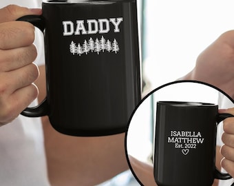 May include: A black ceramic mug with the word "DADDY" in white block letters above a row of white pine trees. A smaller mug in a circle shows the text "ISABELLA MATTHEW Est. 2022" with a heart symbol.