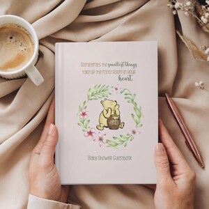 Op de afbeelding: Een roze baby shower gastenboek met een illustratie van Winnie de Poeh. De quote "Sometimes the smallest things take up the most room in your heart." staat op de voorkant gedrukt. De illustratie toont Winnie de Poeh met een pot honing.