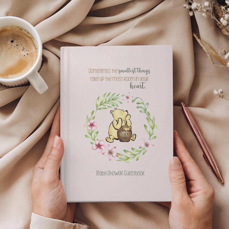 Op de afbeelding: Een roze baby shower gastenboek met een illustratie van Winnie de Poeh. De quote "Sometimes the smallest things take up the most room in your heart." staat op de voorkant gedrukt. De illustratie toont Winnie de Poeh met een pot honing.