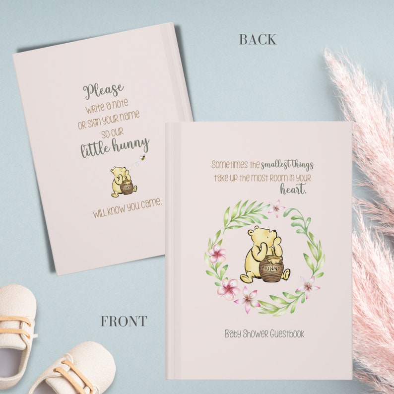 Op de afbeelding: Een roze baby shower gastenboek met een Winnie de Poeh-illustratie. De voorkant heeft de tekst "Schrijf alstublieft een berichtje of teken je naam, zodat onze kleine honingbij weet dat je er was." De achterkant heeft de tekst "Soms nemen de kleinste dingen de meeste ruimte in je hart in."