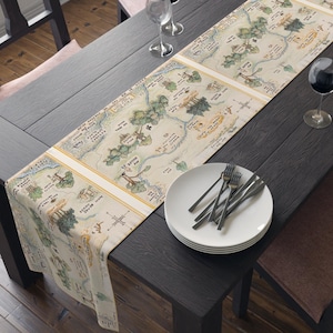 May include: A beige table runner with a vintage map design featuring a forest and the words "The Hundred Acre Wood". The runner is laid on a dark wood table with a white plate and silverware.