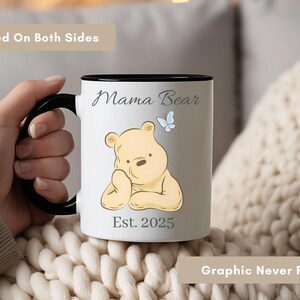 May include: White ceramic mug with a black handle and rim. The mug features a cartoon bear illustration, the words "Mama Bear" and "Est. 2025". A blue butterfly is also included in the design. The text "Printed On Both Sides" and "Graphic Never Fades" are also visible.