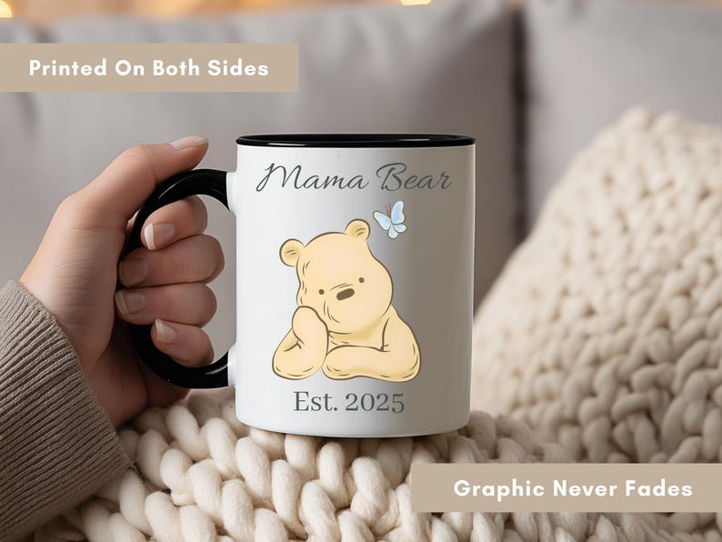 May include: White ceramic mug with a black handle and rim. The mug features a cartoon bear illustration, the words "Mama Bear" and "Est. 2025". A blue butterfly is also included in the design. The text "Printed On Both Sides" and "Graphic Never Fades" are also visible.