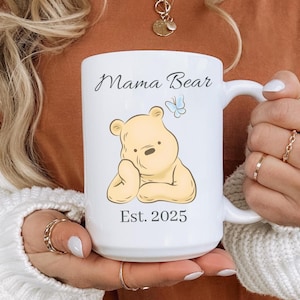 May include: White ceramic mug with the text "Mama Bear" and "Est. 2025". The mug features a cartoon illustration of a yellow bear and a blue butterfly. The mug has a large handle.