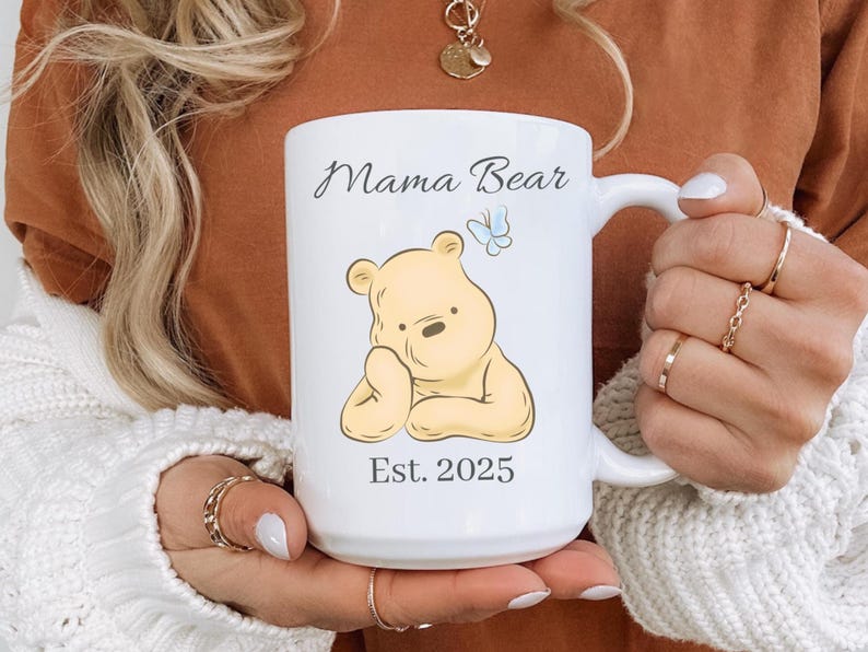 May include: White ceramic mug with the text "Mama Bear" and "Est. 2025". The mug features a cartoon illustration of a yellow bear and a blue butterfly. The mug has a large handle.