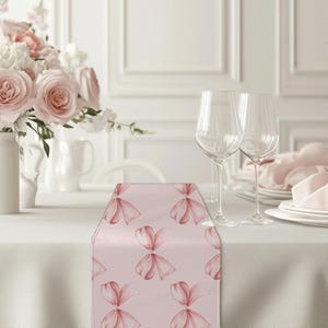 May include: A pink table runner with a repeating pattern of pink watercolor bows. The runner is on a white tablecloth with two wine glasses and a plate on the table.