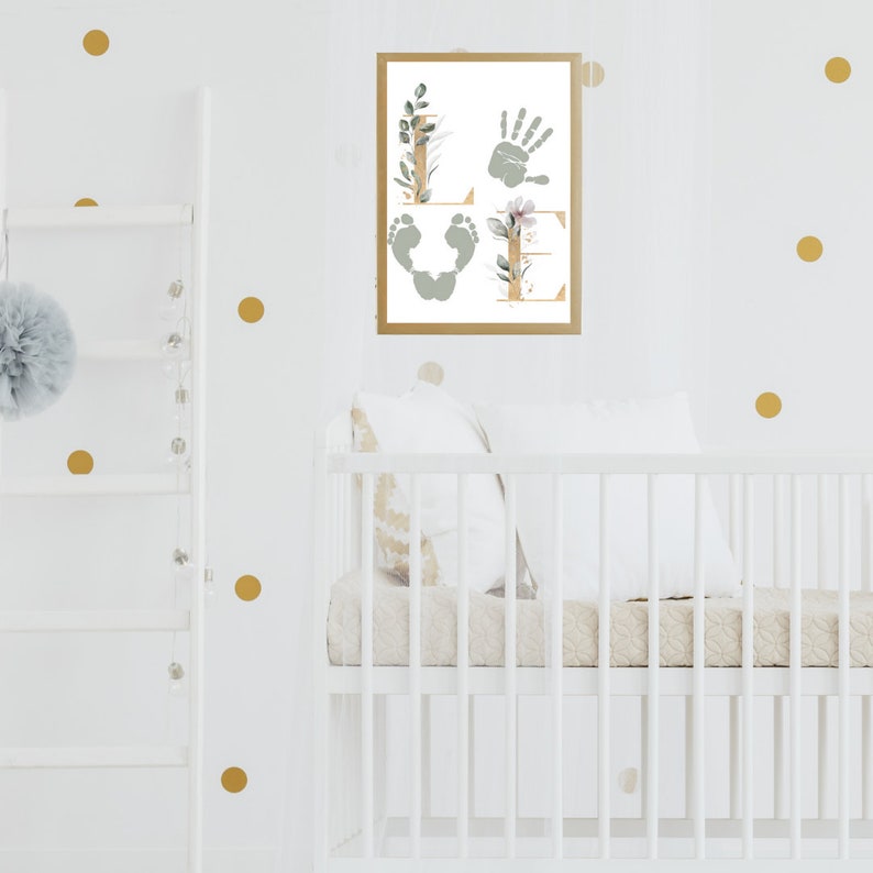 May include: A white crib with a white and beige quilted mattress and a white pillow. A framed print with the word "LOVE" spelled out with handprints and footprints is hanging on the wall behind the crib. The wall is decorated with gold polka dots.