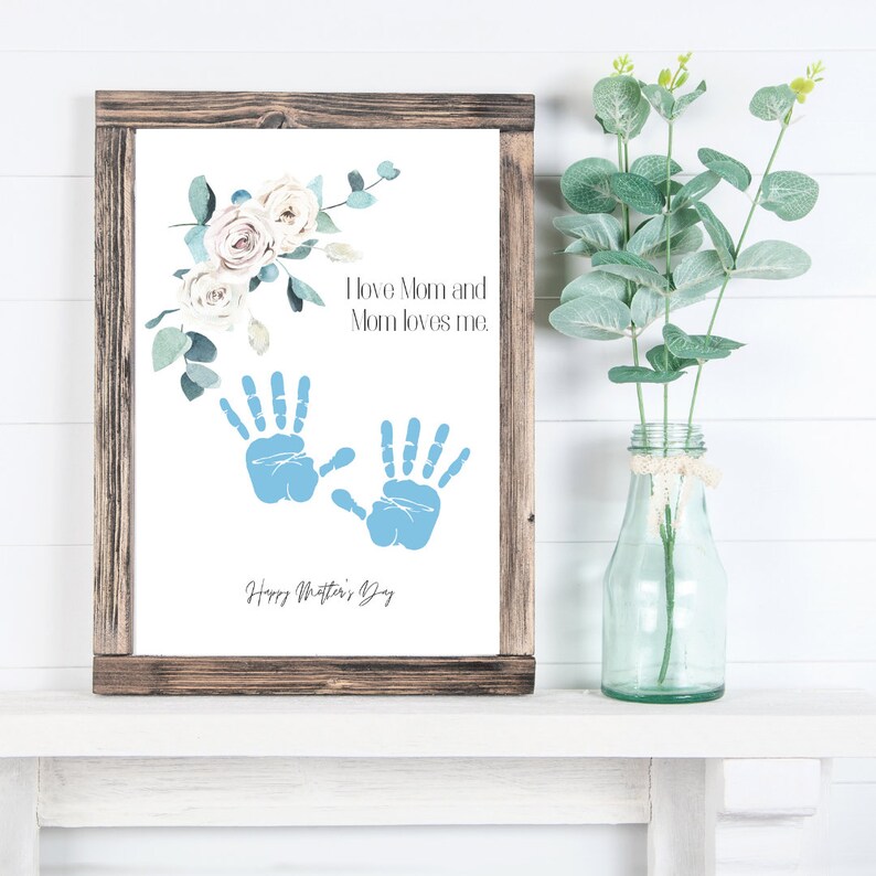 May include: A white framed print with a floral design and blue handprints. The text reads "I love Mom and Mom loves me. Happy Mother's Day."