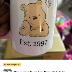 May include: White ceramic mug with a pink handle featuring a cartoon bear illustration and the words "Mama Bear" and "Est. 1997". A butterfly graphic is also present. The mug is a personalized gift.