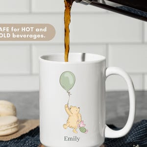 May include: White ceramic mug with a cartoon illustration of a bear holding a green balloon, with a small pig character. The mug has the name "Emily" printed on it. The text "SAFE for HOT and COLD beverages" is also visible.