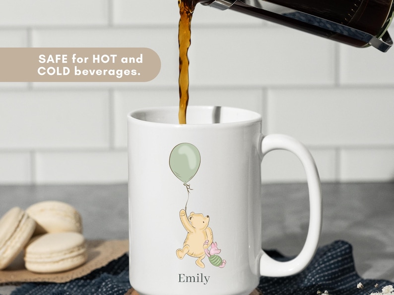 May include: White ceramic mug with a cartoon illustration of a bear holding a green balloon, with a small pig character. The mug has the name "Emily" printed on it. The text "SAFE for HOT and COLD beverages" is also visible.