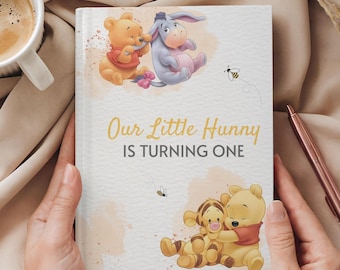 May include: A white journal with a Winnie the Pooh and Piglet illustration. The journal has the text "Our Little Hunny Is Turning One" printed on the front.