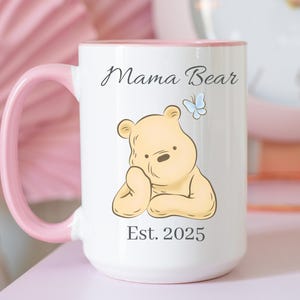 May include: White ceramic mug with a pink handle and interior. The mug features a cartoon bear illustration, a blue butterfly, and the text "Mama Bear" and "Est. 2025".