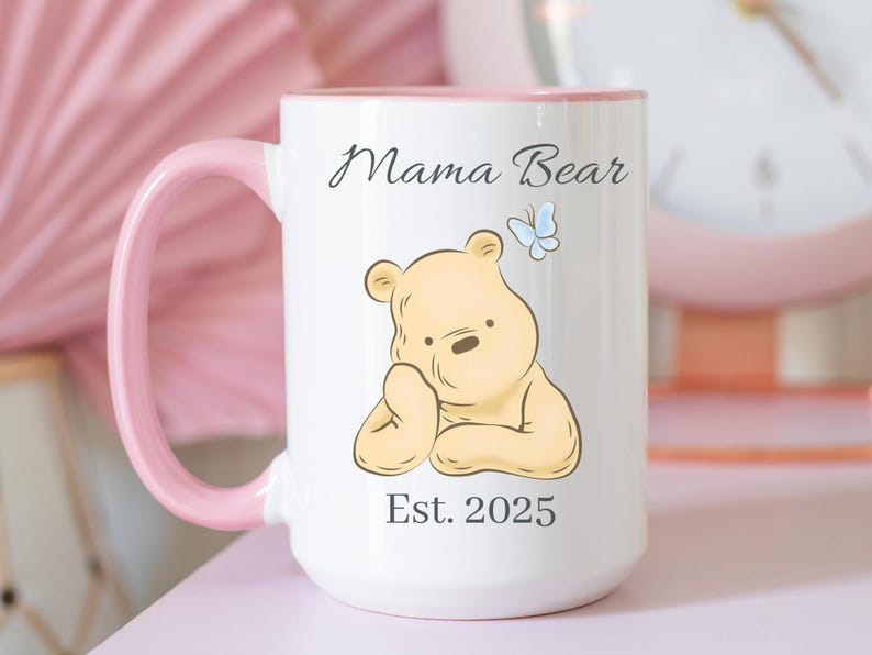 May include: White ceramic mug with a pink handle and interior. The mug features a cartoon bear illustration, a blue butterfly, and the text "Mama Bear" and "Est. 2025".