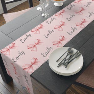 May include: Pink table runner with a repeating pattern of pink bows and the name "Emily" printed in black script.