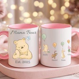 May include: Two white ceramic mugs with pink handles and interiors. One mug features a Winnie the Pooh illustration with the text "Mama Bear" and "Est. 2025". The other mug has illustrations of Pooh characters with balloons and names.