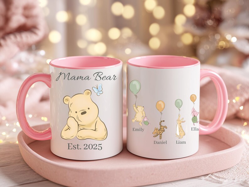 May include: Two white ceramic mugs with pink handles and interiors. One mug features a Winnie the Pooh illustration with the text "Mama Bear" and "Est. 2025". The other mug has illustrations of Pooh characters with balloons and names.