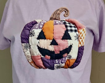 Op de afbeelding: Een lichtpaars t-shirt met een patchwork pompoen ontwerp. De pompoen heeft een lachend gezicht en is gemaakt van verschillende stofpatronen, waaronder oranje, roze, paars en zwart-wit geruit. De pompoen heeft een bruine steel en spinnenweb details.