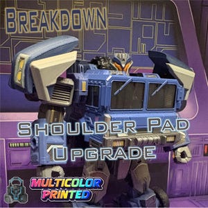 Pode incluir: Um brinquedo robô azul e cinza com o texto "BREAKDOWN SHOULDER PAD UPGRADE MULTICOLOR PRINTED". O brinquedo tem um corpo semelhante a um caminhão e grandes ombreiras. O robô tem detalhes em laranja e preto.