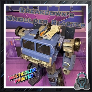 May include: A blue and gray toy robot with the text "BREAKDOWN'S SHOULDER BLASTER" and "MULTICOLOR PRINTED". The toy has wheels and a detailed design, with orange and silver accents. The background is purple.