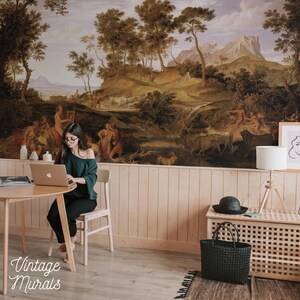 Op de afbeelding: Een kamer met een grote vintage muurschildering die een klassiek landschap met figuren, bomen en bergen uitbeeldt. De kamer heeft een ronde houten tafel, een laptop, een stoel, een lamp en een zwarte draagtas. De tekst "Vintage Murals" staat linksonder.