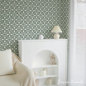 May include: A room interior featuring a green and white patterned wallpaper. A white shelf unit holds a lamp, decorative objects, and a small sculpture. A cream-colored sofa and window with blinds are also visible.