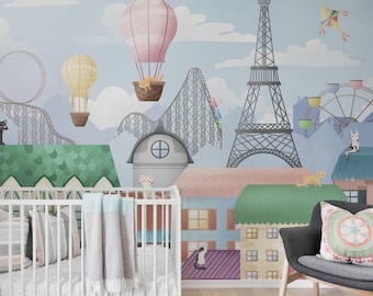 May include: A whimsical nursery scene featuring a mural with a cityscape, hot air balloons, roller coaster, and Eiffel Tower. A white crib with pillows and a blanket is in the foreground. A gray chair and pouf are also visible.