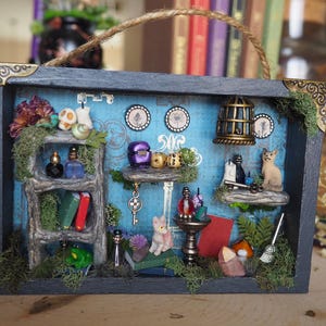 May include: A miniature diorama in a dark gray frame with a blue background. The scene includes a ladder, books, bottles, a birdcage, cats, and other small decorative items. The frame has gold corner accents and a rope handle.