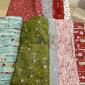 May include: A collection of Christmas-themed fabric rolls. Patterns include red with white script, green with houses and trees, and blue with holiday phrases. Other fabrics feature festive designs in red, white, and gray.